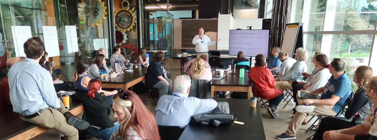 LIPS 2023 workshop participants sit at lab tables in a glass-walled classroom. The workshop leader has a microphone and is referring to a slide on a large monitor.